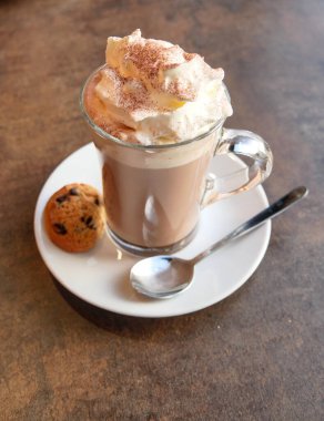Viennese hot chocolate in a transparent cup, placed on a small plate with a cookie and a spoon.