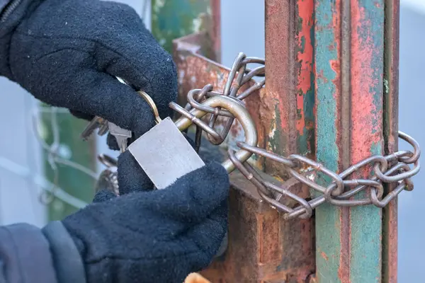 Gloved hands, locking a chain with a padlock, to secure the opening of an old gate with damaged paintwork.