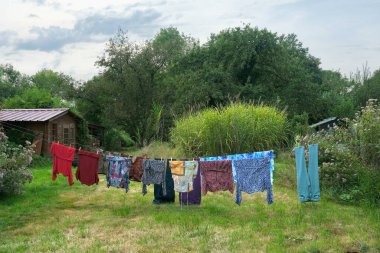 Büyük yeşil bir bahçede uzun bir çamaşır ipinde birkaç renkli bluz kuruyor..