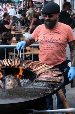 Saint Amand les Eaux, Fransa - 09 08 2024: Sakallı adam, merkezi odun ateşiyle büyük yuvarlak bir barbeküde bir dizi sosis pişiriyor..