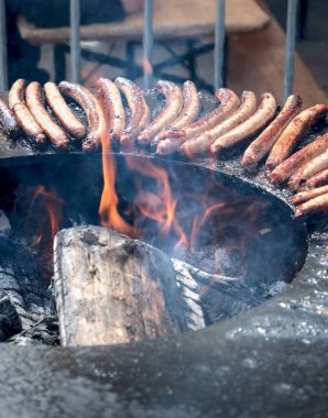Büyük dairesel bir barbeküde pişen bir dizi sosis ve merkezi bir ateş püskürtüsü..