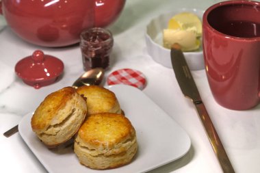 Küçük kare bir tabakta çörekler, bir kavanoz reçel, tereyağı, çaydanlık ve bir kupa çayla çevrili. Öğleden sonra çayının tüm unsurları