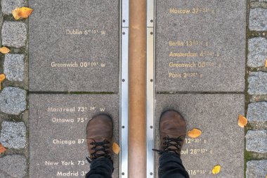 Ayakları Greenwich, Londra 'da, küresel zaman dilimlerini temsil eden yere oyulmuş boylam işaretleri ile Birincil Meridyen üzerinde duruyor..