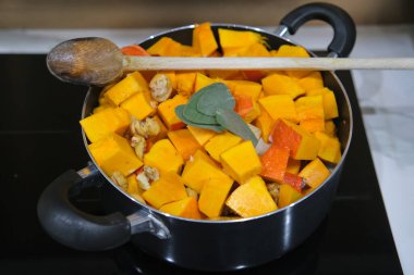 Yeni doğranmış tereyağlı kabak, kestane ve adaçayı siyah bir tencerede, ahşap bir kaşıkla modern bir fırında pişirmek için hazırlanıyor..