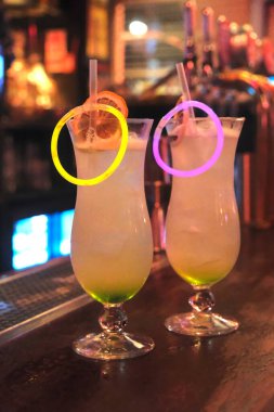 Close-up of two vibrant cocktails with neon glow sticks and dried orange garnish, placed on a wooden bar counter under warm bar lighting. clipart