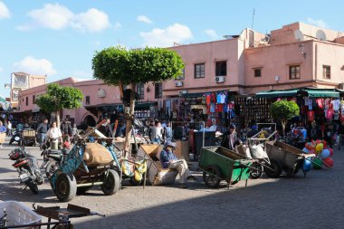 Marrakech ,Morocco - 12 30 2024 : A bustling Moroccan market square with vendors, carts, and pedestrians, surrounded by pinkish buildings selling clothes, souvenirs, and local goods. clipart