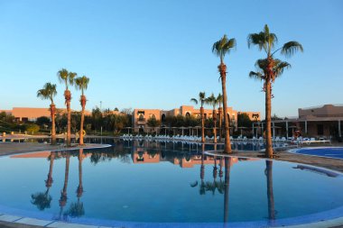 Parlak mavi gökyüzünün altında, güneş pansiyonları ve terracotta renkli binalarla çevrili, berrak bir havuza yansıyan yüksek palmiye ağaçlarıyla güzel bir tatil yeri..
