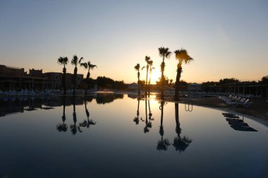 Gün batımında sakin bir tatil köyü sahnesi. Palmiye ağaçları ve onların yansımaları durgun bir havuzda, altın ışıkla yıkanmış, huzurlu ve rüya gibi bir atmosfer yaratıyor..