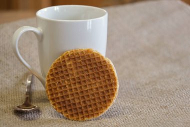 Yanı başında karamel waffle kurabiyesi olan beyaz bir fincanın yakın çekimi, yanında bir gül altın kaşığı, desenli bir çuval bezi arka planda..