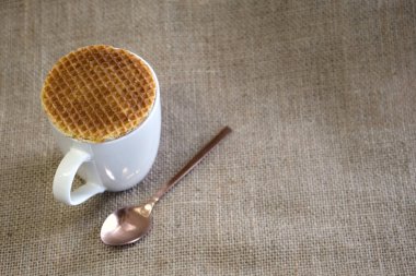 A close-up of a white mug with a caramel waffle cookie resting on its top, accompanied by a rose gold spoon, set on a textured burlap background. clipart