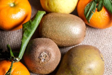 Yapraklı armutlar, kiviler ve mandalinalar da dahil olmak üzere taze meyvelerin tepeden tırnağa görüntüsü doğal ışık altında desenli bir çuval bezi üzerine dizilmiştir..