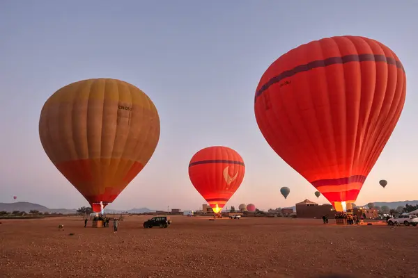 Marakeş, Morocoo - 01 02 2025: 3 sıcak hava balonu şafak vakti bir çöl manzarasında uçuşa hazırlanıyor. Gökyüzü açık ve balonlar ocaklarla aydınlatılıyor.