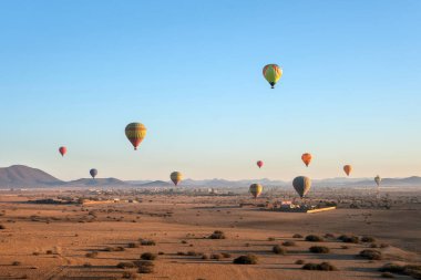 Sıcak hava balonları güneş doğarken engin bir çöl manzarasının üzerinde yüzer ve altın kumun üzerine uzun gölgeler bırakır. Açık gökyüzü ve uzak dağlar derinlik katar.
