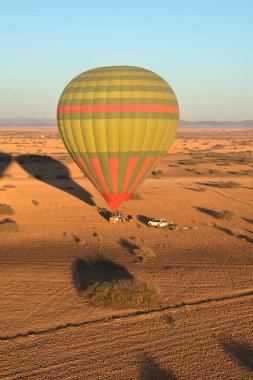 Kırmızı ve yeşil çizgili renkli bir sıcak hava balonu, açık mavi gökyüzünün altında, park halindeki beyaz bir aracın yanında, bir çöl manzarasında duruyor..