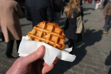Bir el, arka planda bulanık yayaların olduğu hareketli bir açık hava ortamında eğlenmeye hazır, peçeteye sarılı altın, çıtır Belçika waffle 'ını tutuyor..