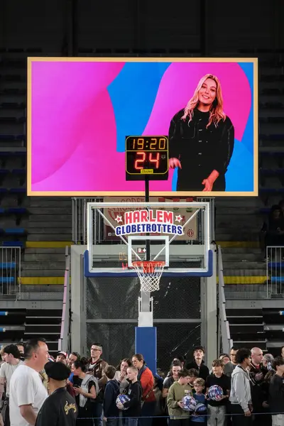 Orchies, Fransa - 03 28 2025: Harlem Globetrotters basketbol potası içinde canlı bir logo, saati ve skoru okuyan bir skor tahtası ve basketbol topu tutan coşkulu bir kalabalık.