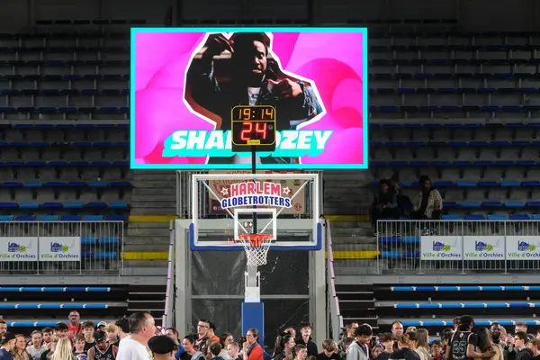 Orchies, Fransa - 03 28 2025: Harlem Globetrotters basketbol potası içinde canlı bir logo, saati ve skoru okuyan bir skor tahtası ve basketbol topu tutan coşkulu bir kalabalık.