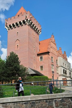 Poznan, Polonya - 04 / 22 / 2025: Poznan Şatosu, yüksek kulesi, kırmızı çatısı ve mavi gökyüzü altında manzaralı yeşilliği olan tarihi tuğla kale, Polonya.
