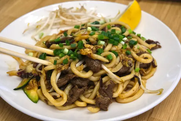 Bir tabak kızarmış udon eriştesi sığır eti, yeşil soğan, fasulye filizi ve limon dilimi ile süslenmiş. Çubuklar eriştenin üzerinde dinleniyor..