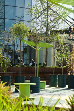Poznan, Polonya - 04 25 2025: Canlı yeşil masalarla açık hava kafe sahnesi ve çiçek yetiştiren genç ağaçlar arasında sandalyeler, yumuşak gün ışığı altında modern cam bir binaya karşı hazırlanıyor.