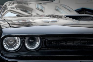 Pürüzsüz farları ve ızgarası olan parlak siyah arabanın ön tarafı modern, mimari bir sahnede kentsel çevreyi yansıtıyor..