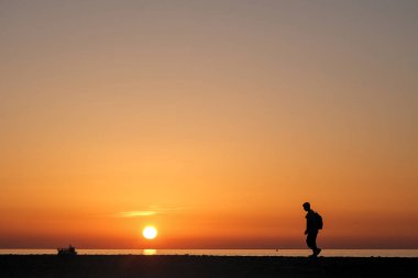 Gün batımında sakin bir plajda tek başına yürüyen biri. Canlı turuncu gökyüzü, yumuşak dalgalar ve dingin ruh hali huzuru ve yalnızlığı çağrıştırıyor..