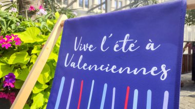 Valenciennes, Fransa - 08 05 05 2025: Valenciennes 'de yaz titreşimleri renkli güverte koltuğu, palmiye ağaçları ve parlak mavi gökyüzü altında tarihi bir bina.