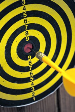 Sarı dart tahtaya saplanmış siyah ve sarı hedef tahtasına isabet ediyor. Tahta duvara monte edilmiş, doğruluğu, odaklanmayı ve başarıyı simgeliyor..