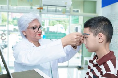 Orta yaşlı göz doktoru genç bir çocuğu muayene ediyor ve Asyalı bir çocuğa göz muayenesi yapıyor.