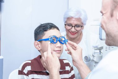 Gözlüklü gülümseyen çocuk modern klinikte, doktorlarda ve göz hekimliği kliniğindeki hastaları desteklemek için büyükannesiyle birlikte pediatrik göz doktorunu muayene ediyor..