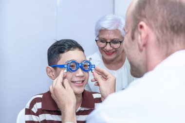 Gözlüklü gülümseyen çocuk modern klinikte, doktorlarda ve göz hekimliği kliniğindeki hastaları desteklemek için büyükannesiyle birlikte pediatrik göz doktorunu muayene ediyor..