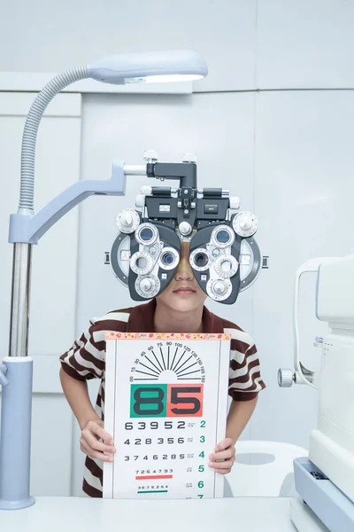 Asian boy looking through optical phoropter during eye exam, diagnostic ophthalmology equipment, selective focus