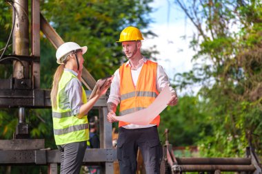 Hard Hats 'deki erkek ve kadın endüstriyel mühendisler dizüstü bilgisayar kullanırken yeni projeleri tartışıyorlar. Ağır bir endüstriyel fabrikada çalışırken jestler yapıyorlar..