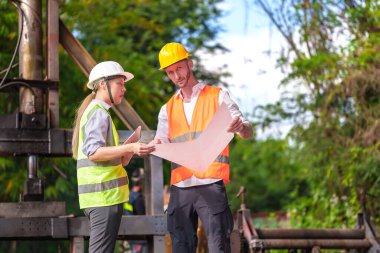 Hard Hats 'deki erkek ve kadın endüstriyel mühendisler dizüstü bilgisayar kullanırken yeni projeleri tartışıyorlar. Ağır bir endüstriyel fabrikada çalışırken jestler yapıyorlar..