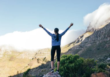 Genç, zayıf bir kadının, bir patika koşusu sırasında bulutlu bir dağın önünde ellerini kaldırışının arka görüntüsü. Tam boy bir sporcu kadın kayanın üzerinde duruyor ve dağlara bakıyor..