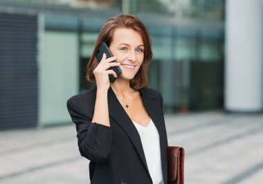 Resmi giysiler içinde gülümseyen bir kadın ofis binasının önünde beklerken telefonla konuşuyor. Orta yaşlı bir kadının portresi gözlerini kaçırıyor ve cep telefonuyla konuşuyor..
