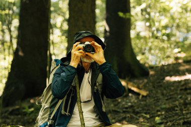 Yürüyüş sırasında ormanda oturan şapkalı kıdemli bir turist fotoğraf çekiyor.