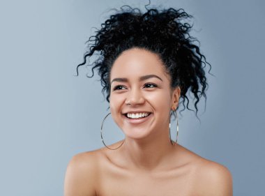 Genç, mutlu, doğal güzelliği ve kıvırcık saçları olan, gri bir arka plana bakan bir kadın.