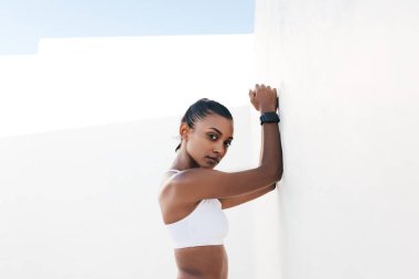 Beyaz duvara eğilip doğrudan kameraya bakan fit bir kadının portresi. Spor yaparken spor yapmak için kısa bir ara veriyor..