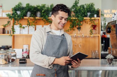 Elinde dijital tablet olan önlüklü bir erkek barista kahve tezgahına yaslanıyor.