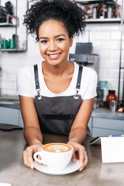 Smiling waitress with curly hair with a freshly prepared cappuccino at the counter 