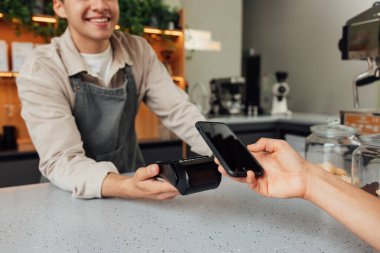 Tanımlanamayan kafe sahibi tezgahta kart makinesi tutarken, müşteri akıllı telefondan ödeme yapıyor.