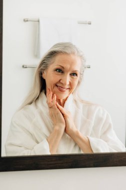 Gülümseyen son sınıf öğrencisi kadın banyo aynasında yansımasına bakıyor.