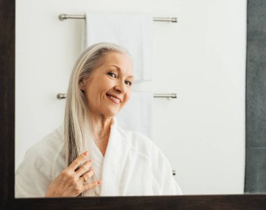 Gülümseyen yaşlı kadın uzun gri saçlarına dokunuyor banyo aynasındaki yansımasına bakıyor.