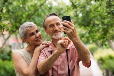 Yaşlı, gülümseyen bir çift parkta selfie çekiyor.