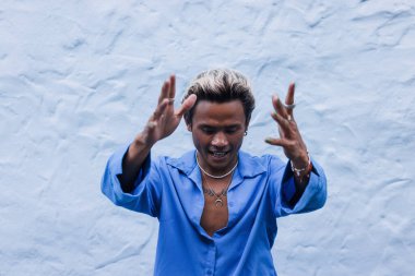 Mavi gömlekli genç dışarıda duvara poz veriyor.