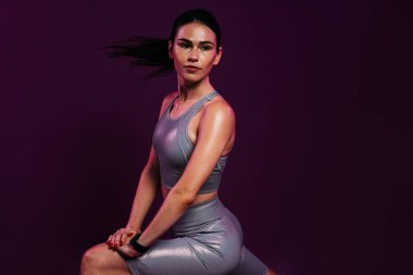 Gümüş rengi spor giysili genç bir kadın, vücudunun alt kısmını morumsu bir arka plana karşı ısıtıyor.