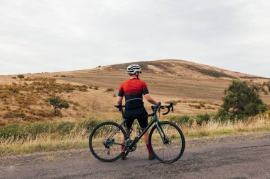 Arkaya bakan kadın bisikletçi boş kır yolunda bisiklete yaslanıyor.