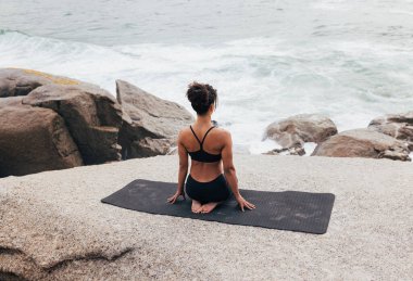 Sıska kadın spor kıyafetleri içinde mindere oturmuş okyanusa bakarken.