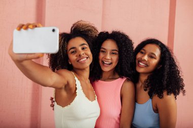Parlak giyinmiş genç kadınlar bir arada durup cep telefonuyla selfie çekiyorlar. Mutlu kadınlar dışarıda pembe duvarlara karşı eğleniyorlar..